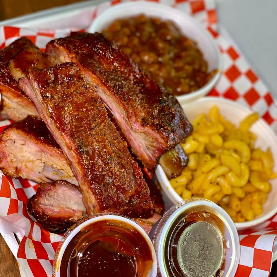 BBQ ribs with mac and cheese, baked beans, and two cups of sauce on red and white checkered paper.