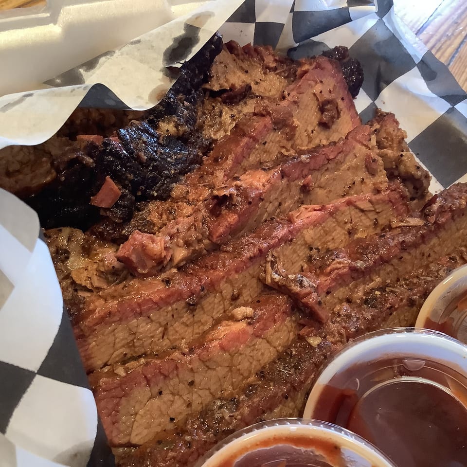 Sliced smoked and grilled meats served on black-and-white checkered paper with small cups of dipping sauces.