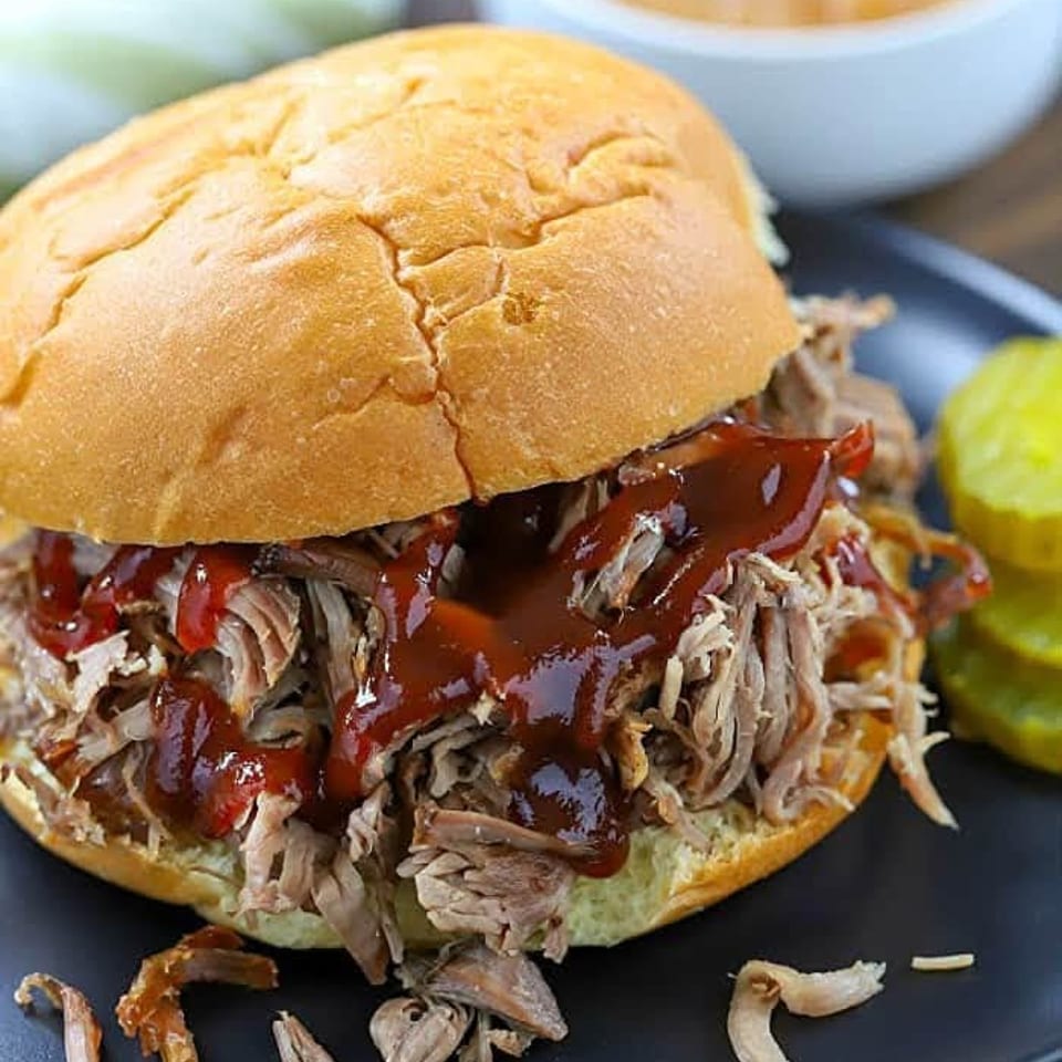 Pulled pork sandwich with barbecue sauce, pickles, and a side of sauce on a black plate.