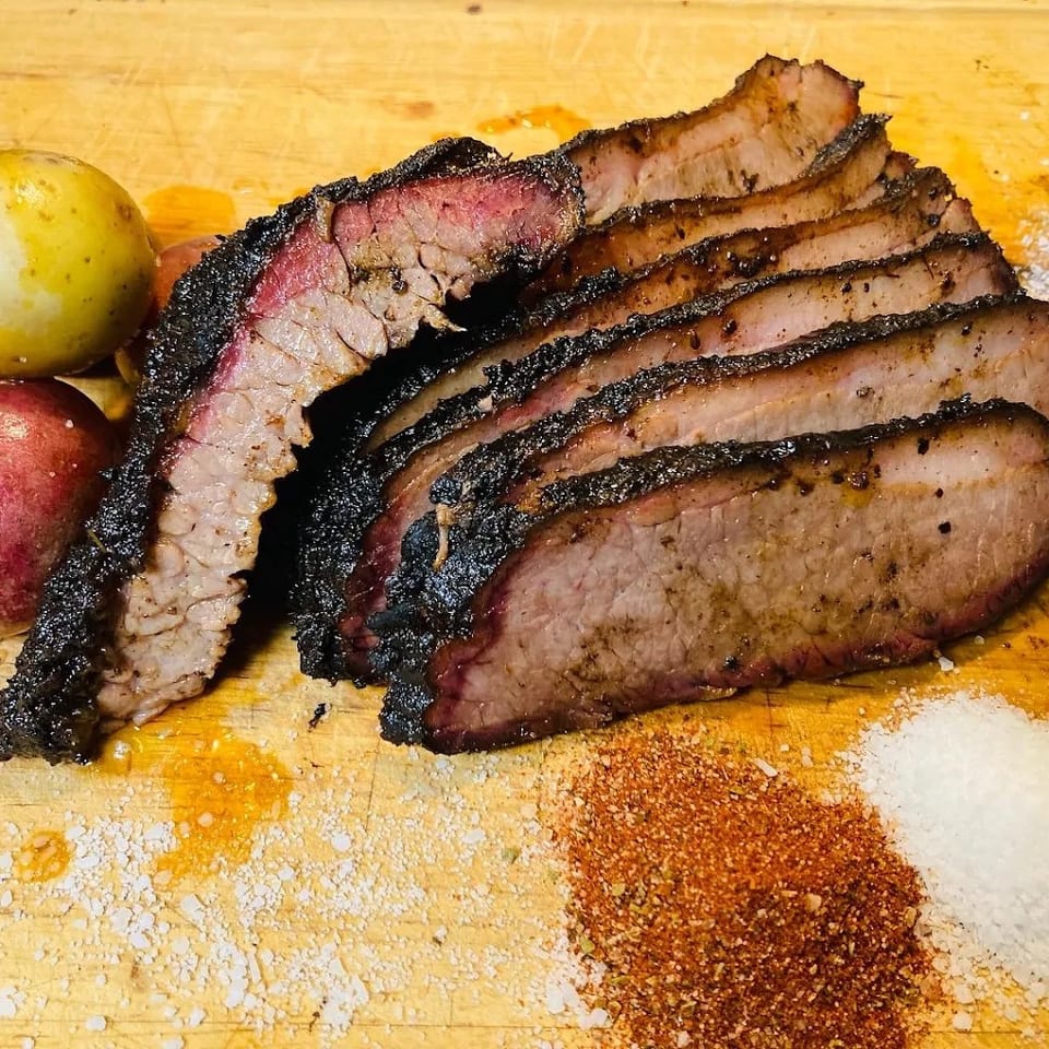 Sliced smoked brisket with a dark crust rests on a wooden cutting board beside red and yellow potatoes and spices.